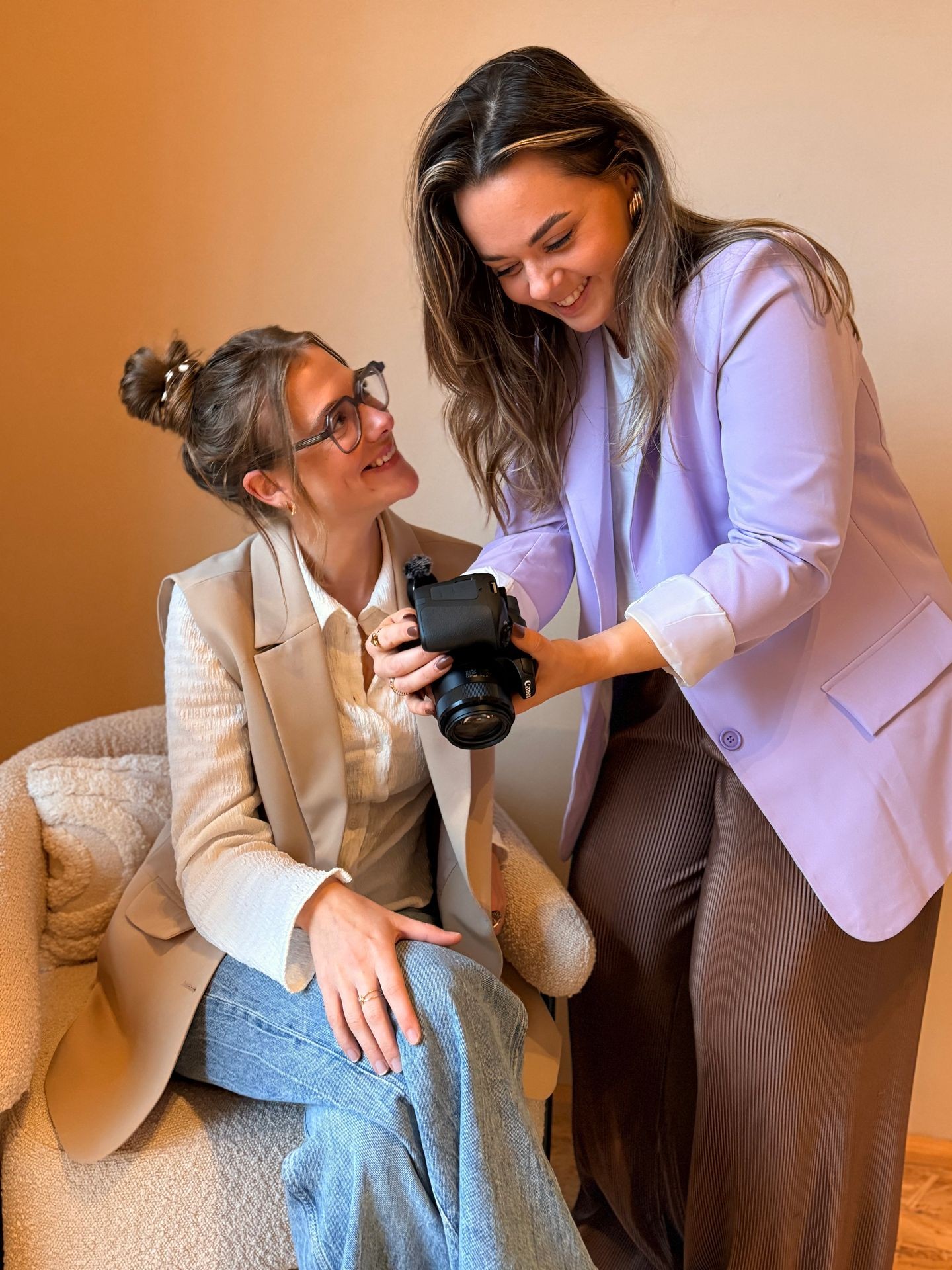 Two women smiling as they look at a camera in a cozy indoor setting.